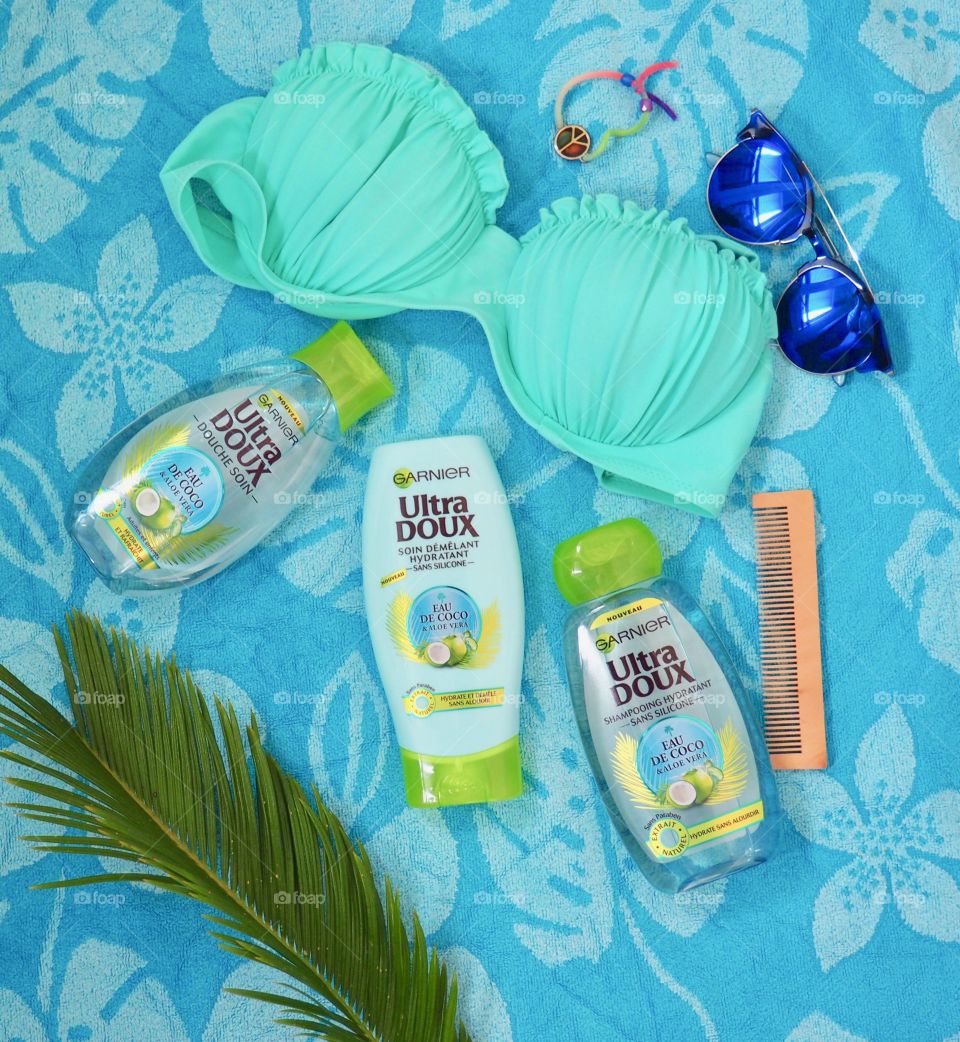 Garnier eau de coco shampoo, conditioner and bath gel on a blue towel with comb and palm leaf and blue sunglasses and bikini.