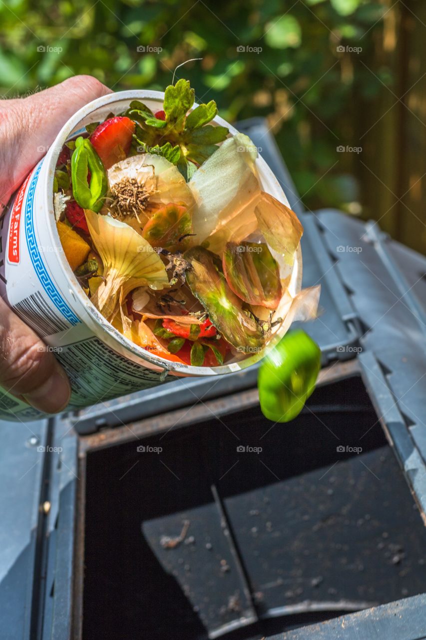 Vertical photo of fruit and vegetable remnants being dumped into a compost tumbler with blurred motion of a pepper top falling