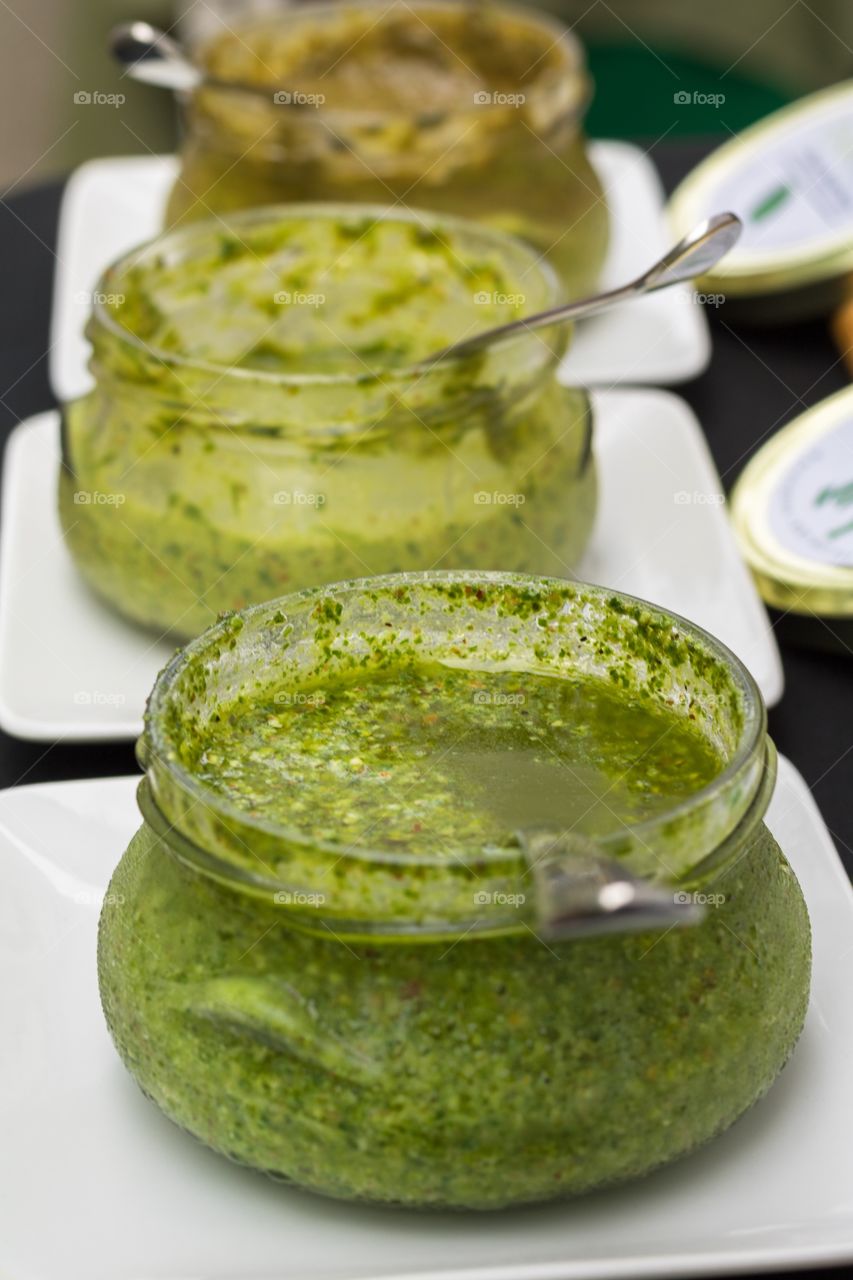 Vertical closeup soft focus photo of 3 pesto variations in glass jars with stainless steel spoons on white serving plates