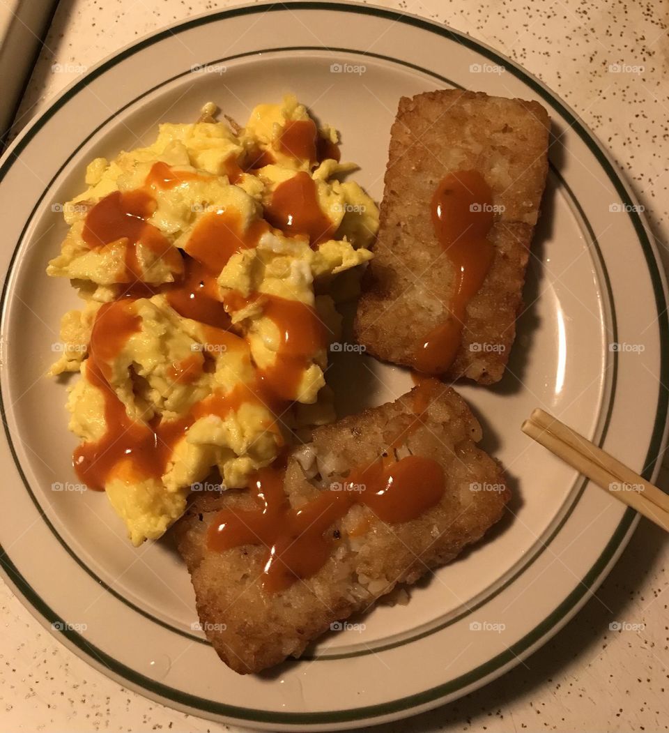 Breakfast for supper at home. This tasty meal is comprised of scrambled eggs and two hash browns. Some wing sauce is added to spice up the food.