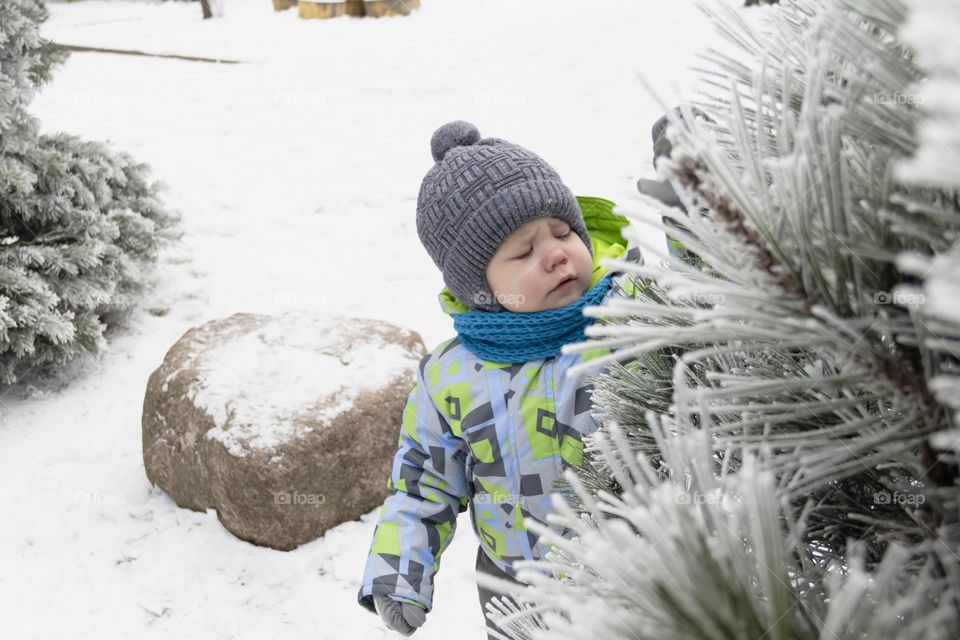 A small, carefree boy walks in winter through the white snow in the park, near the trees in the snow.