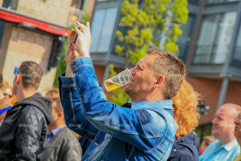 Man taking a photo with his smartphone. With his hands full on his smartphone he's holding his drink in his teeth