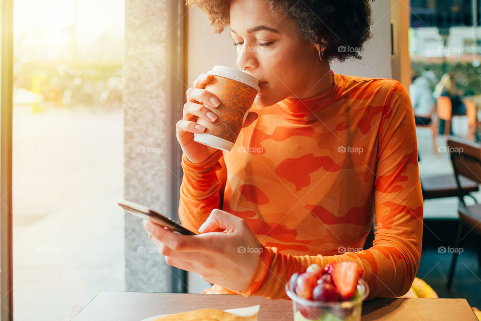 young beautiful black woman sitting at the bar drinking a coffee using smartphone - connection, break, smart working