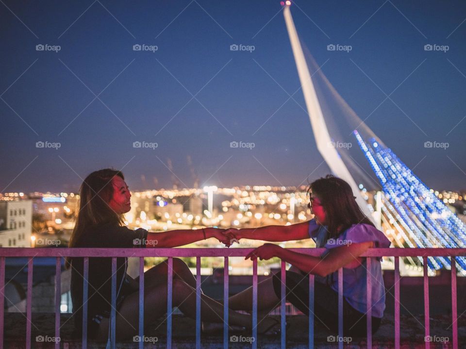 Sisters for life: two young girls are smiling and holding hands on the rooftop of a building under the beauty of the city night scene