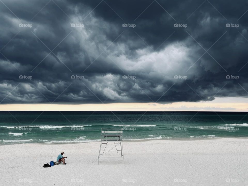 Lone man on beach sitting next to empty metal lifeguard stand. The ominous clouds indicate a gathering thunder storm.