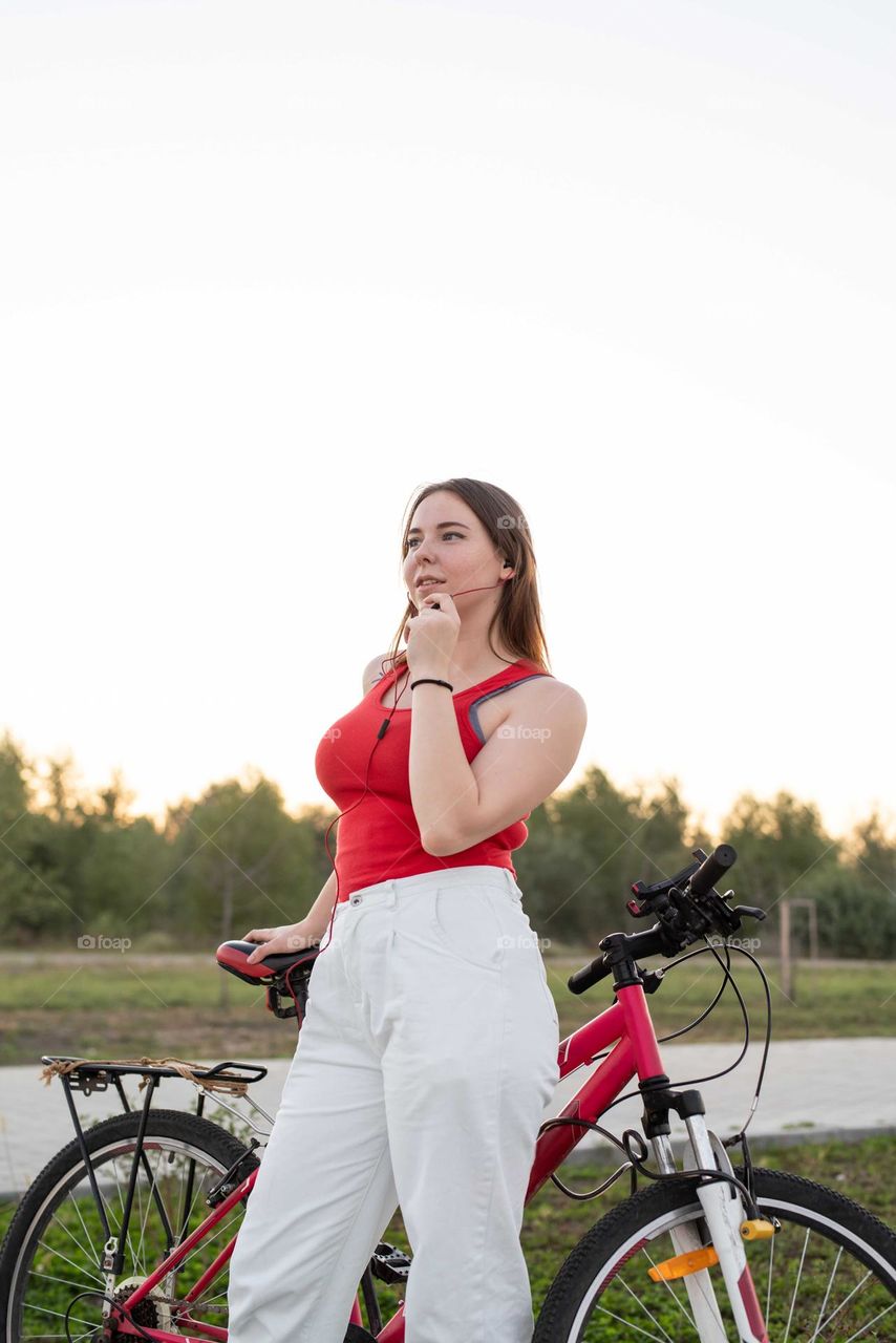 young smiling woman in red shirt and white pants listening to the music standing near her bicycle