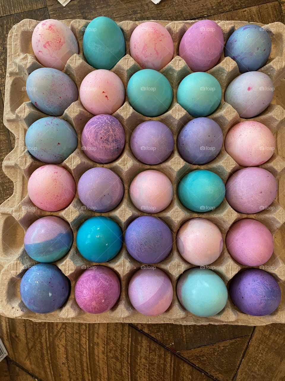 A variety of pastel, dye-colored Easter Eggs drying on an egg carton on the refrigerator shelf. The eggs are pink, purple, green, turquoise, blue, and teal. 