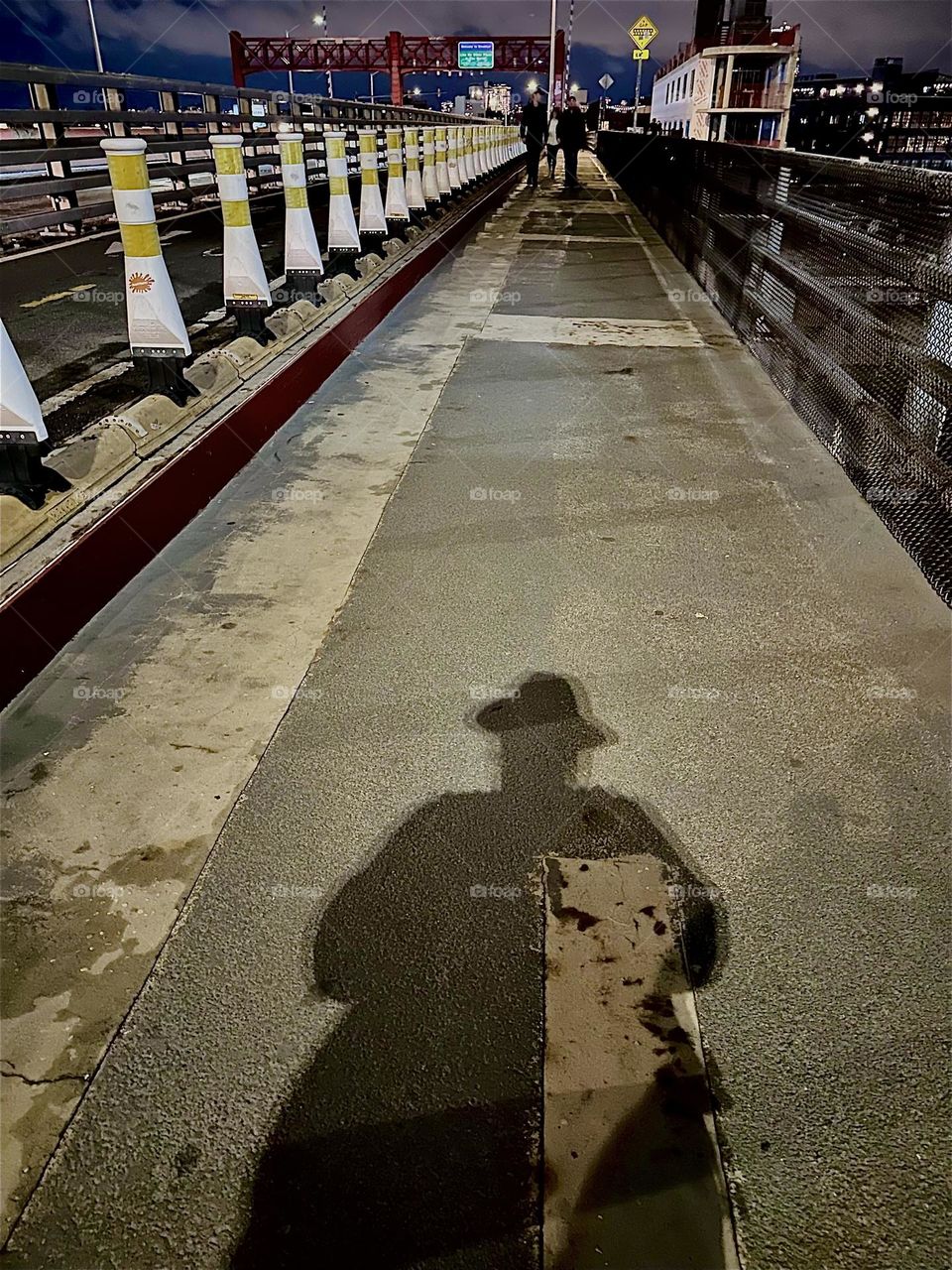 A dramatic shadow is cast onto the pedestrian lane of the “Pulaski Bridge” at “Newtown Creek”, the bridge that connects LIC, Queens to “Greenpoint”, Bklyn which is the direction we are currently facing. 2023. Hypnotic Productions
