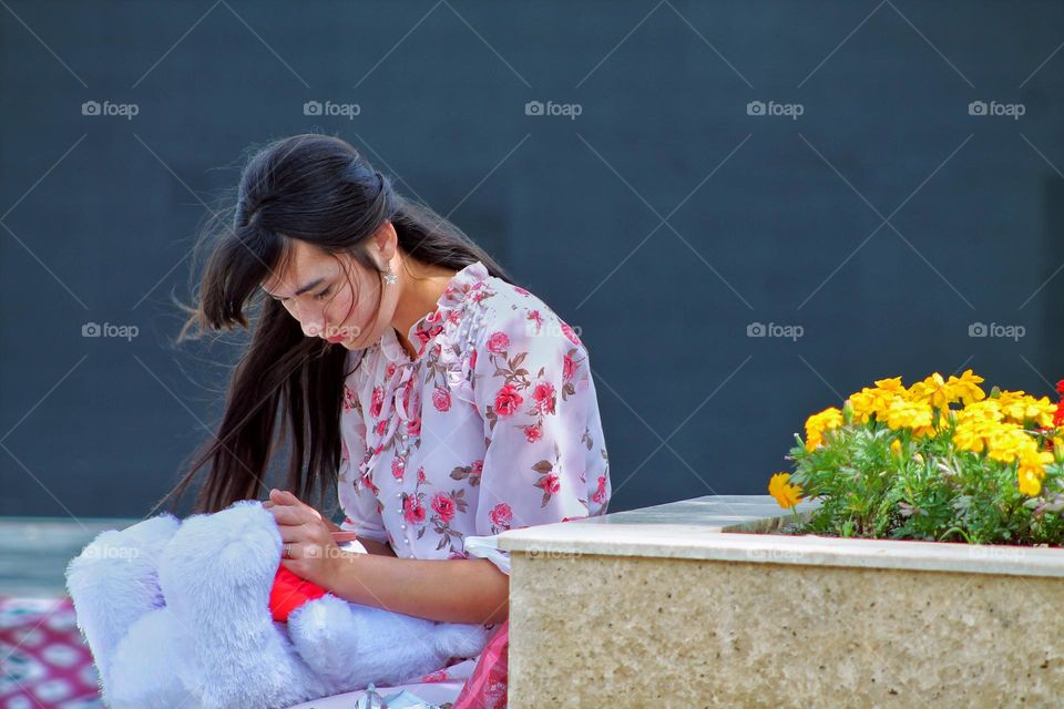 girl with a teddy bear sits near a flowerbed