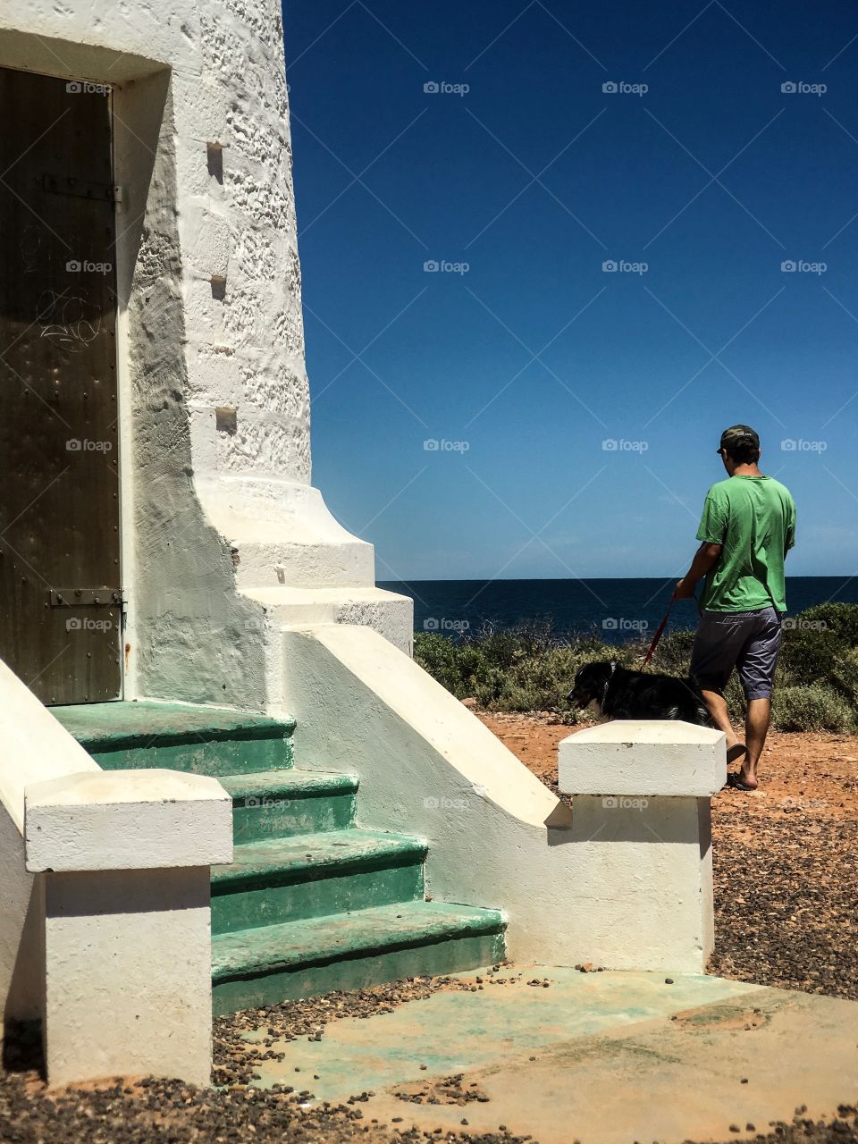 Youth in green tee shirt and shorts walking dog by lighthouse at ocean on clear blue sunny day