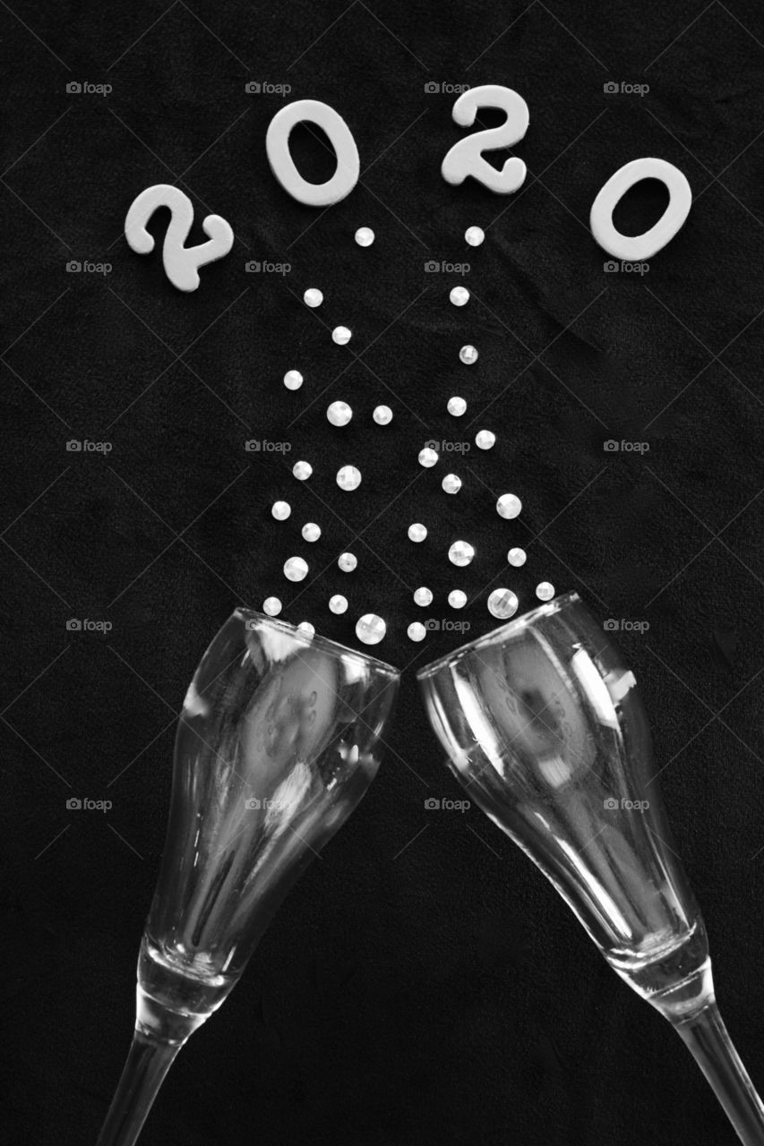 Flat lay of the numbers ‘2020’ above champagne glasses with rhinestone bubbles on black background in black and white - portrait format