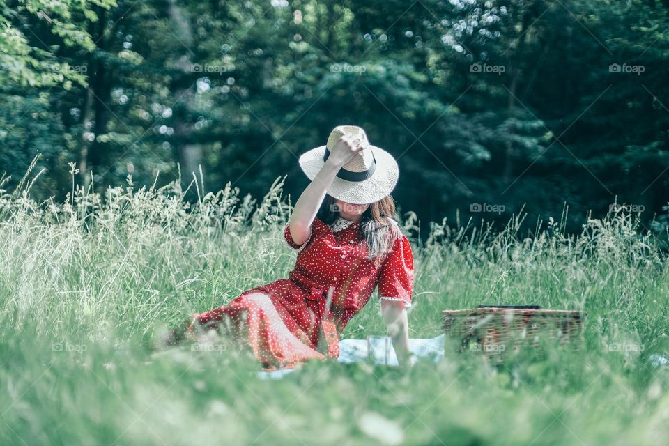 One young caucasian brunette girl in a red dress sits in a meadow with tall grass covering her face from the sun with a straw hat, close-up side view.