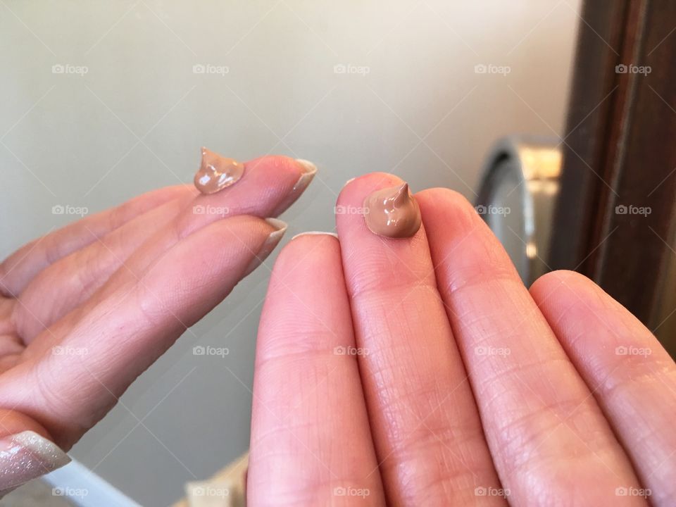 Woman's hand with small amount of tinted moisturizer on finger tips for facial beauty routine reflection in mirror at home 