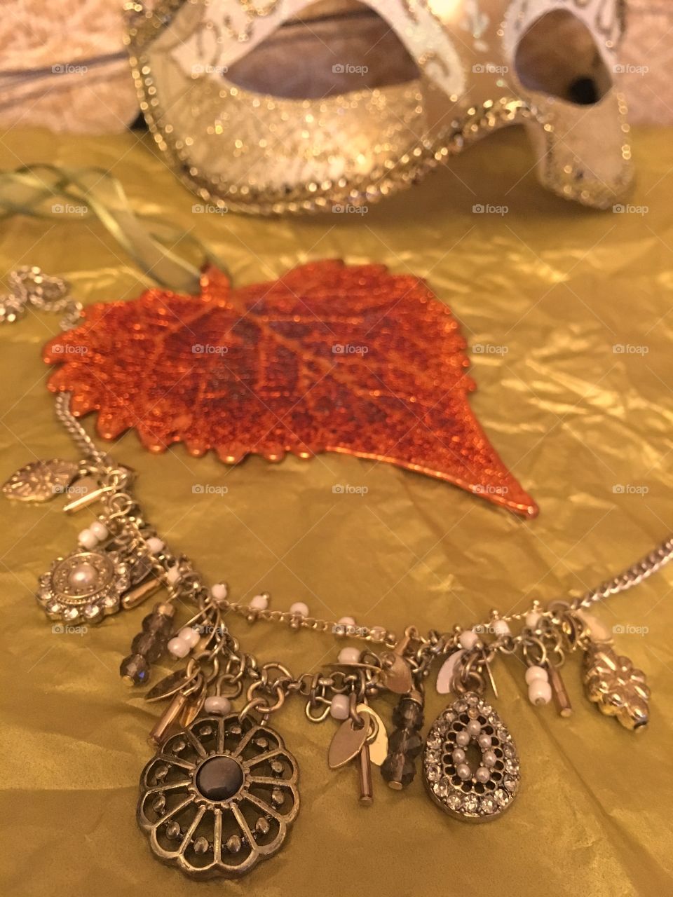Gold charm necklace with a metallised cottonwood leaf decoration with masquerade mask in background 