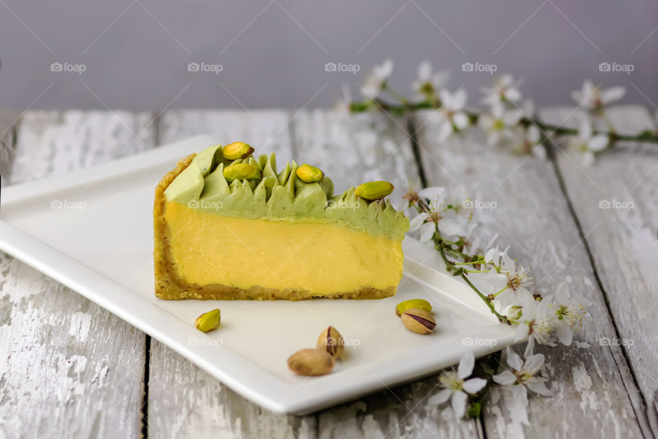 Mango-pistachio cheesecake on white plate at wooden old table and flowered branches.