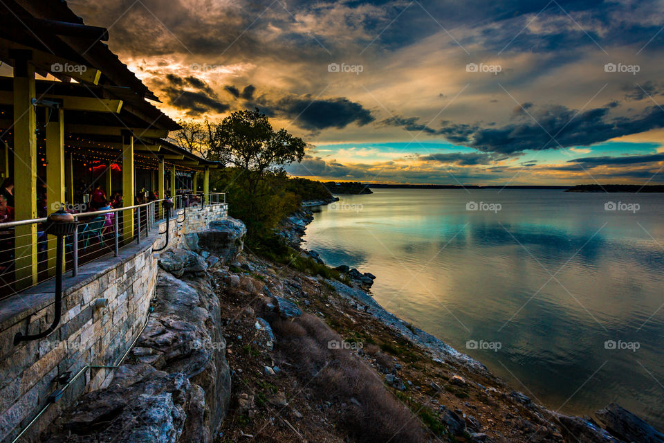The evening sun setting behind the cliffs on a lake behind a restaurant bar setting the sky aglow with orange blue and purple colors in the moody clouds. The vivid colors are reflecting in the Surface of the water.