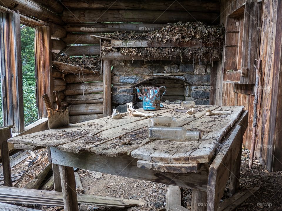 Inside an old abandoned cabin with a huge pack rats nest on the fire place mantle and a old wooden table.