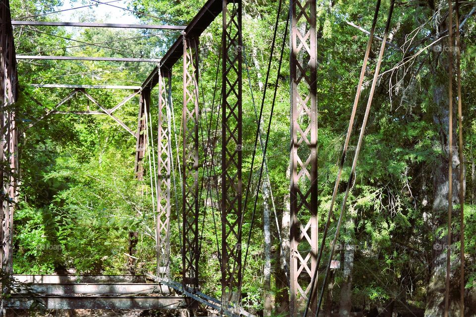 The side of a very old metal bridge that is dilapidated and rusted surrounded by lush green trees
