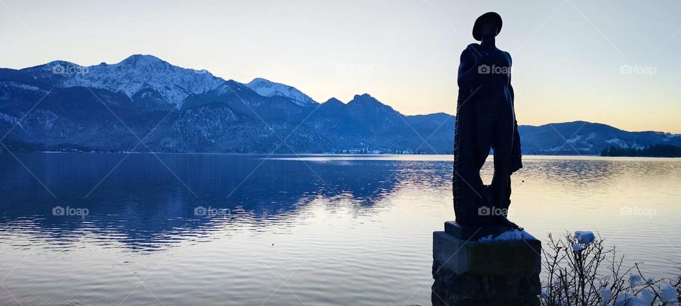 A replica of the “Fisherman” statue by “Richard Miller” adorns “Lake Kochel”, a pre Alpine freshwater lake by the resort town of “Kochel” known amongst other things for its natural hot geyser springs, spas and saunas. 2024. Hypnotic Productions