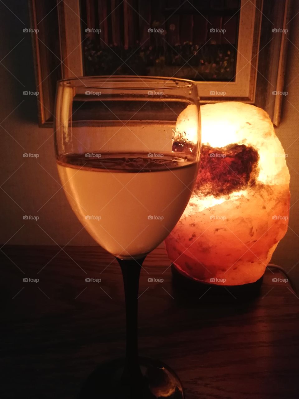 A see-through wine glass with a black leg standing on a brown wooden table in a dark room. The surface of the glass is wet for the cold drink. Clear drops become visible.Behind is an orange light, made of salt and a painting on the wall.