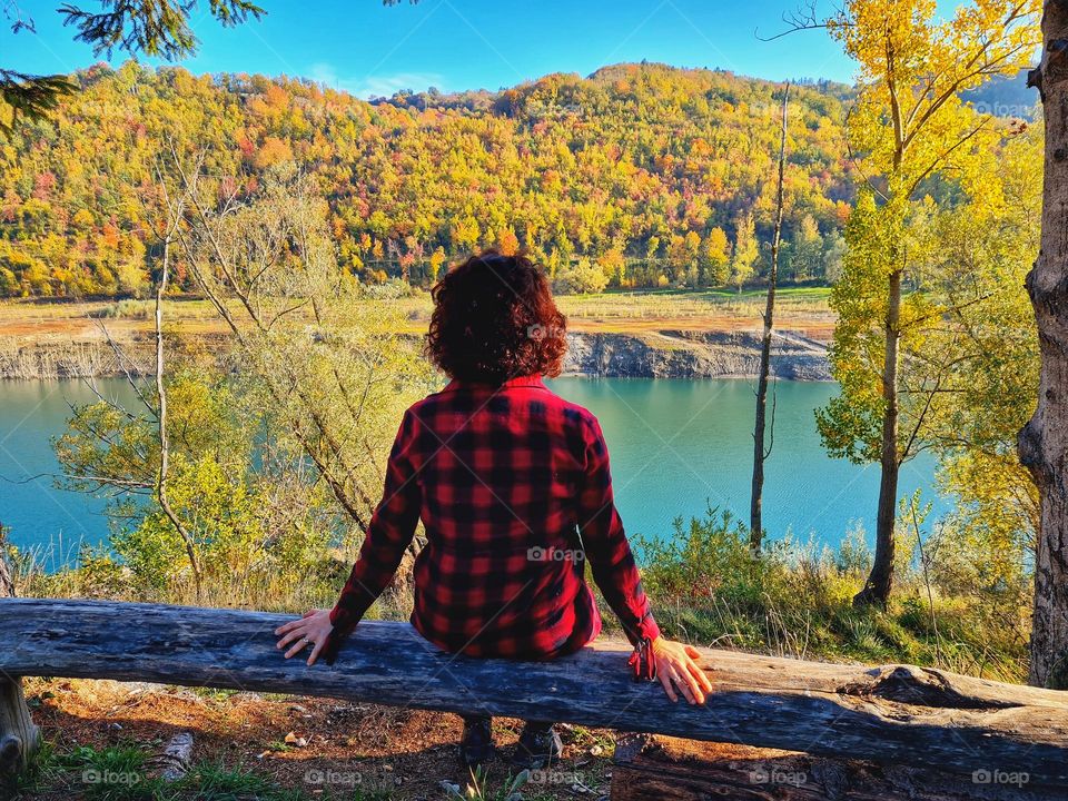 Shoulders hiker with warm checkered flannel shirt sits on a log and admires the lake in autumn