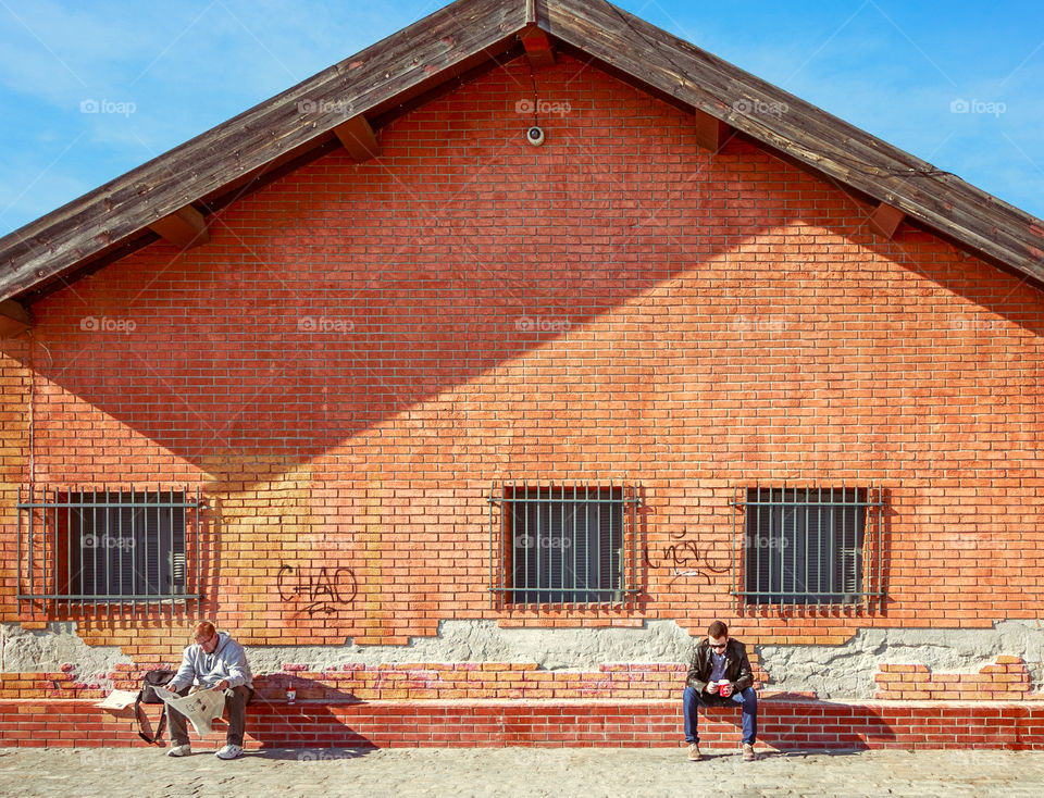 Two Men Sitting Outdoors In Front Of A Building, One Checking His Cellphone And Drinking Coffee And The Other Reading Newspaper. Keeping Social Distancing.
