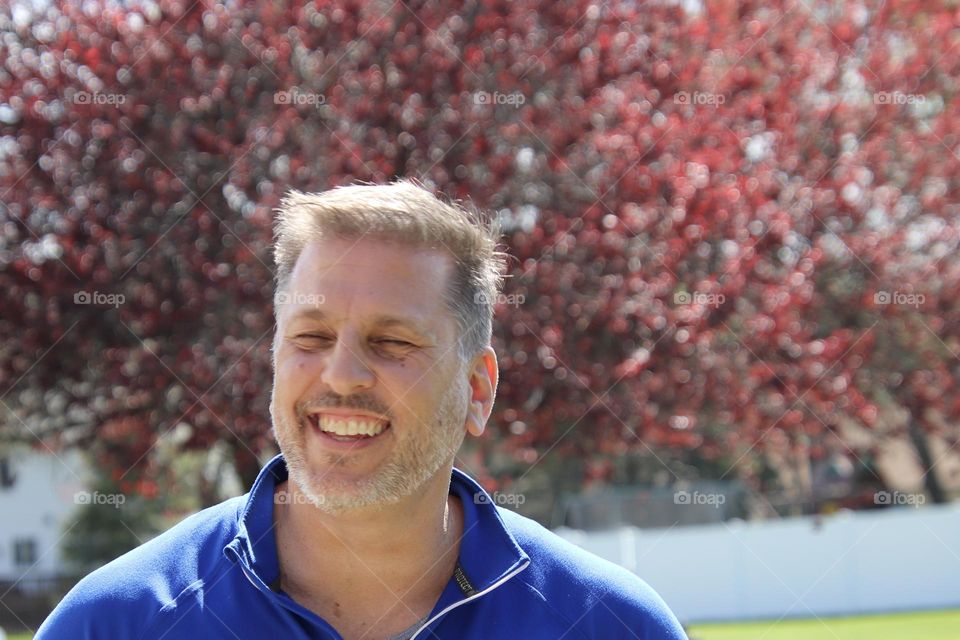 Man with short beard smiling outdoors on sunny day plum tree in background 