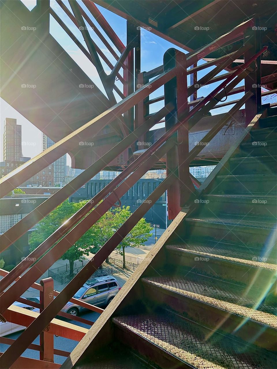 The red metal stairs of the “Pulaski Bridge” at “Newtown Creek” in “Greenpoint”, Bklyn have strong geometry. The angled railings with four parallel bars create interwoven patterns especially when shadows fall across. 2024. Hypnotic Productions 