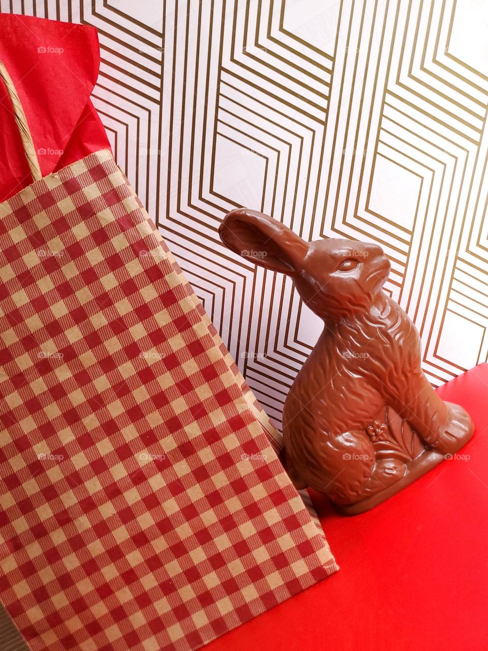Faux chocolate bunny next to a red and light brown plad gift bag with a gold and white multiple diamond shape background and red surface.