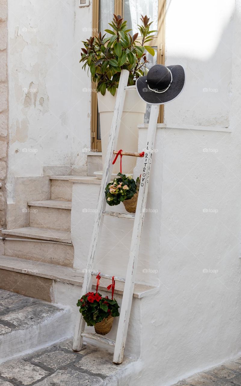 A beautiful view of a decorative staircase with flowers in a pot resting against the wall of a house in the white city of Lstuni, Italy, side view, close-up. Urban plants concept, creative decoration.