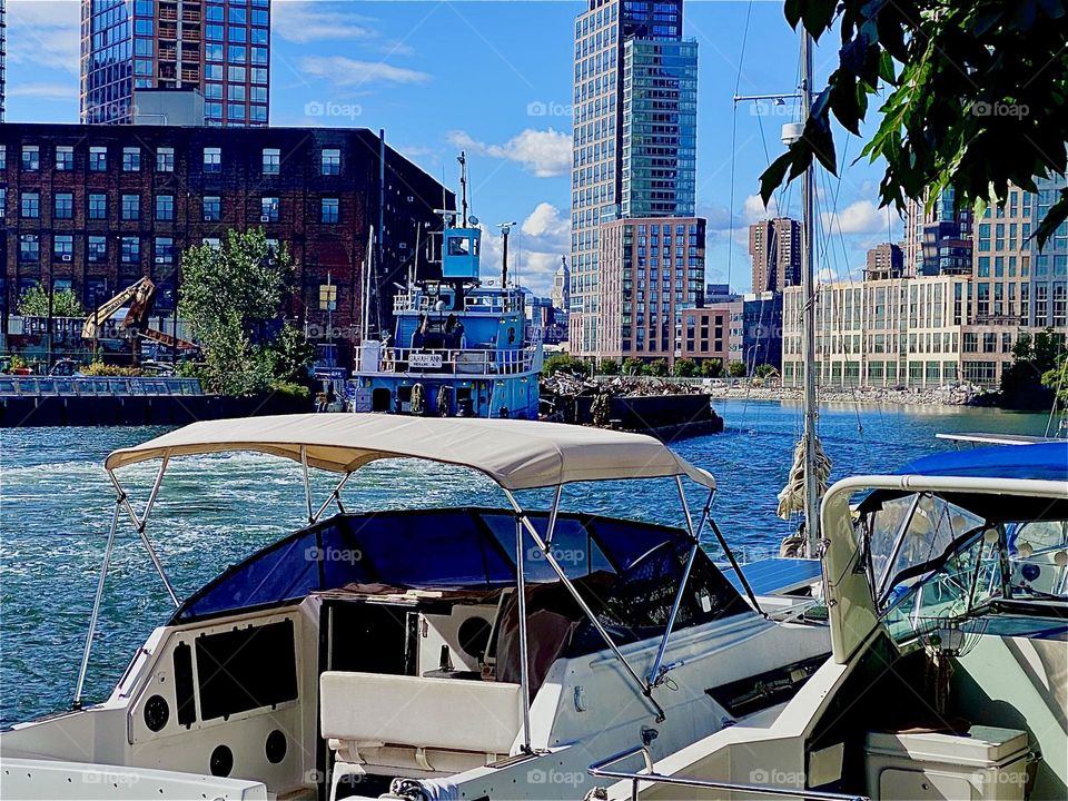 Behind the motorboats that are tied to the shore here a large industrial barge is floating by at „Newtown Creek“ by the „Pulaski Bridge“ In LIC, Queens on a bright sunshiny day in October 2023. Hypnotic Productions
