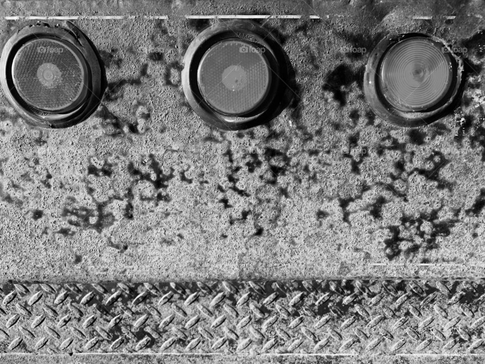 Three rear lights of a a heavy-duty truck on a rusted iron tailgate with diamond-plating in monochrome