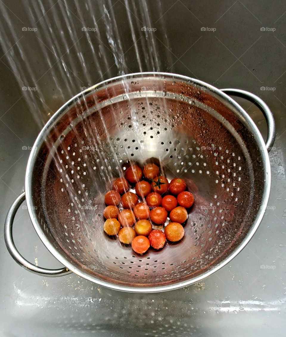 Kitchen sink faucet spraying water into silver metal colander that's holding ripe cherry tomatoes.