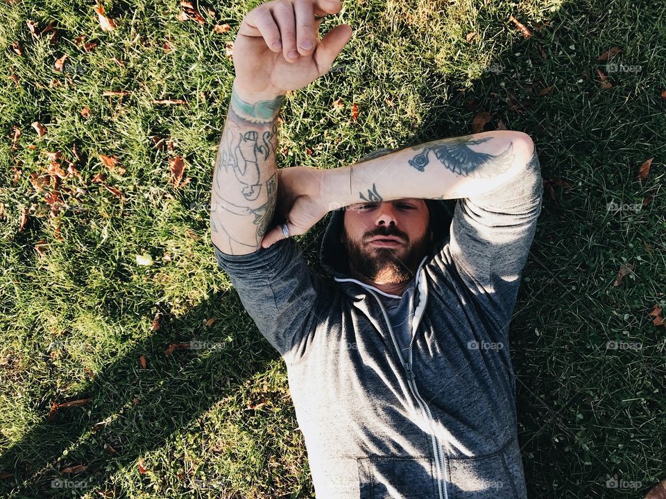 Guy with tattoos laying on the grass, looking up with his arms up. 