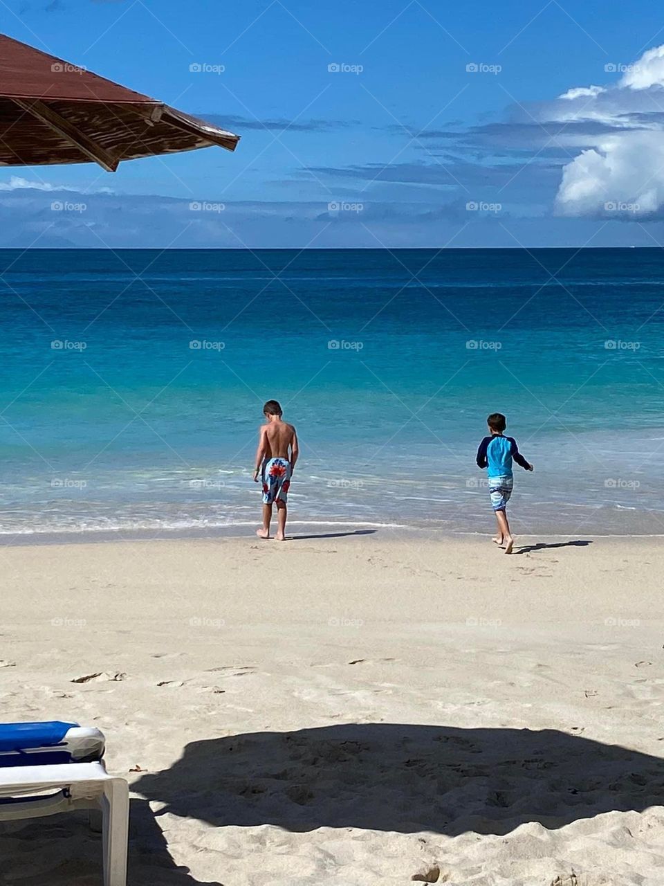 Two children run from the clean sandy beach to the pristine aqua colored ocean. The water ombrés from aquamarine to turquoise, from turquoise to dark blue underneath a light blue sky with a white cloud. 