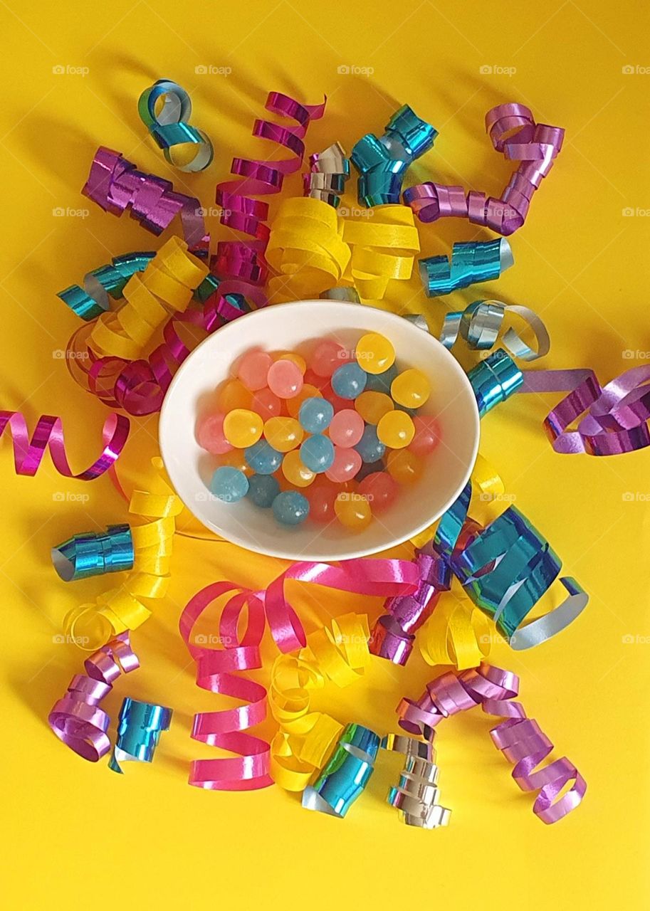 Colourful flatlay on a yellow background composed of pink, purple, yellow, blue and silver gift package ribbons with a little china-bone bowl full of yellow, pink and blue Haribo jelly beans