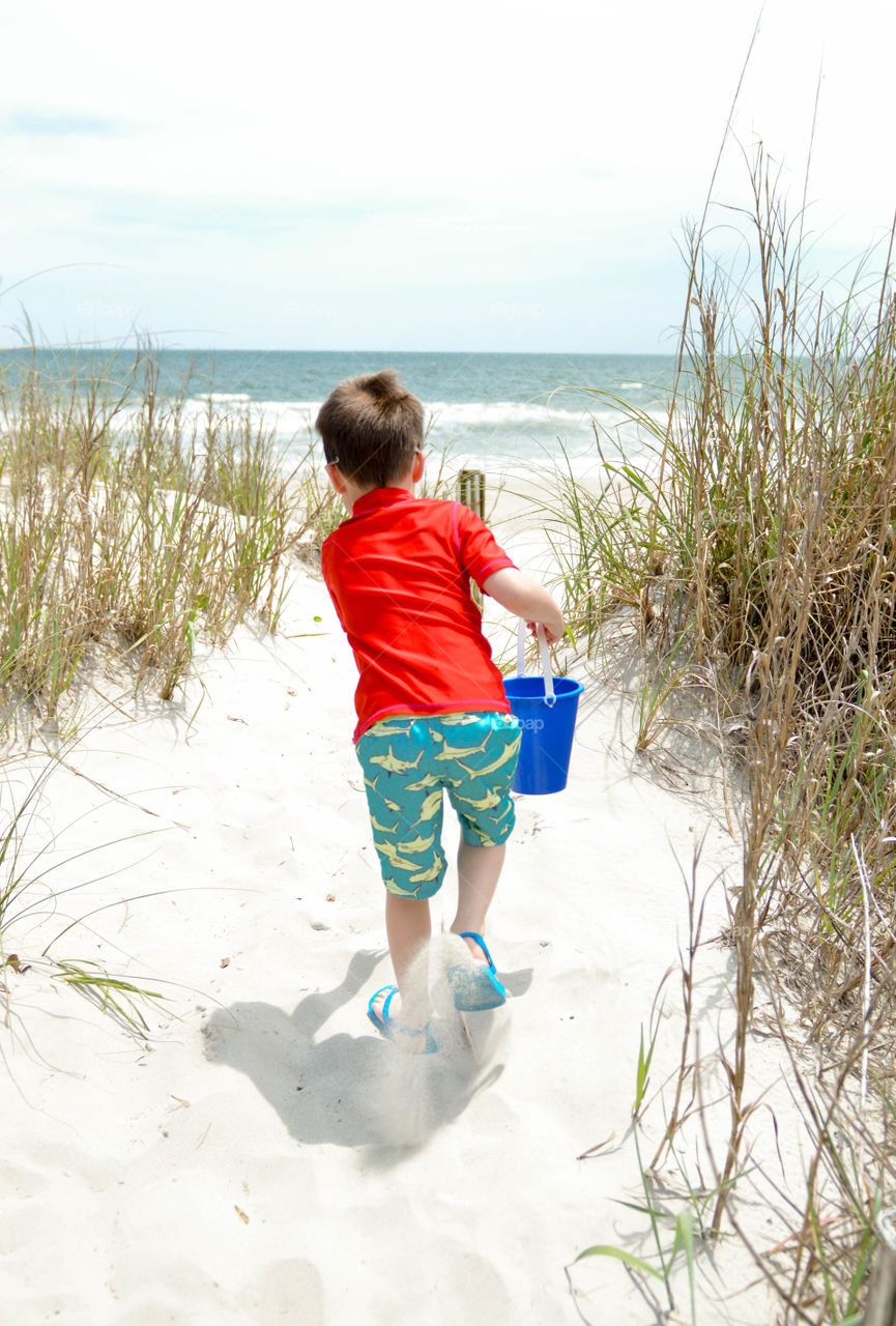 Young boy running through a beach access sand dune to the ocean with a pail