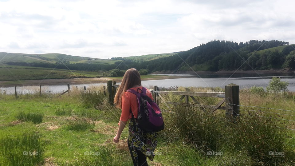 Long Haired Girl Wearing A Dress And A Rucksack Walking Through Grass With Hills And Lake In The Background - Goyt Valley Peak District National Park - Beautiful Landscape