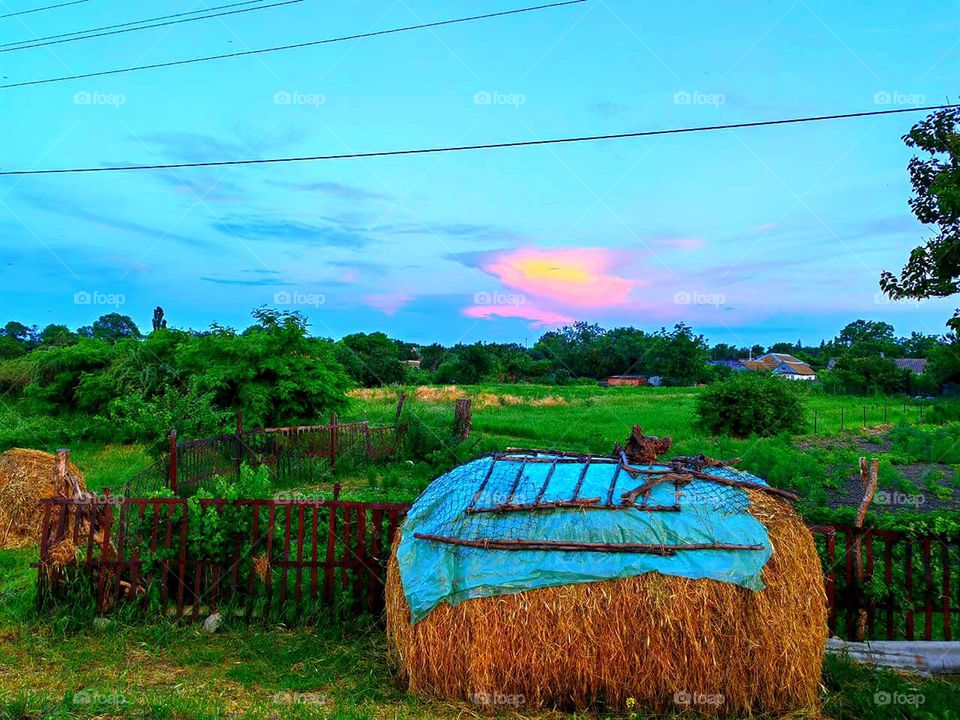 Countryside. Haystack next to a wooden wicker fence. Houses are visible in the distance. Green grass and trees. On the horizon, a blue sky with a pink patch of clouds from the sunset