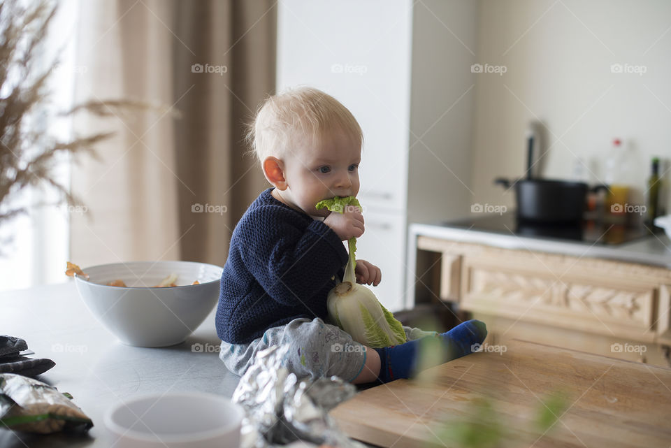 Cute baby boy sits on a table in the kitchen and eats a green salad leaf