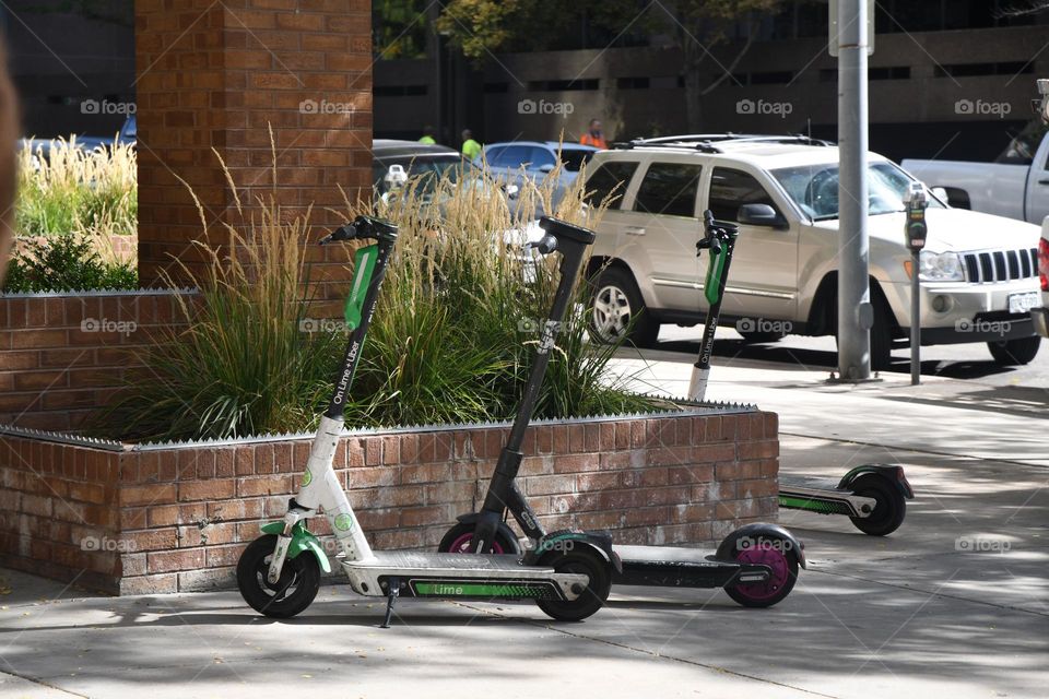 Three electric pay by the hour scooters are parked on a sidewalk in the downtown metropolitan district of Denver, Colorado.
