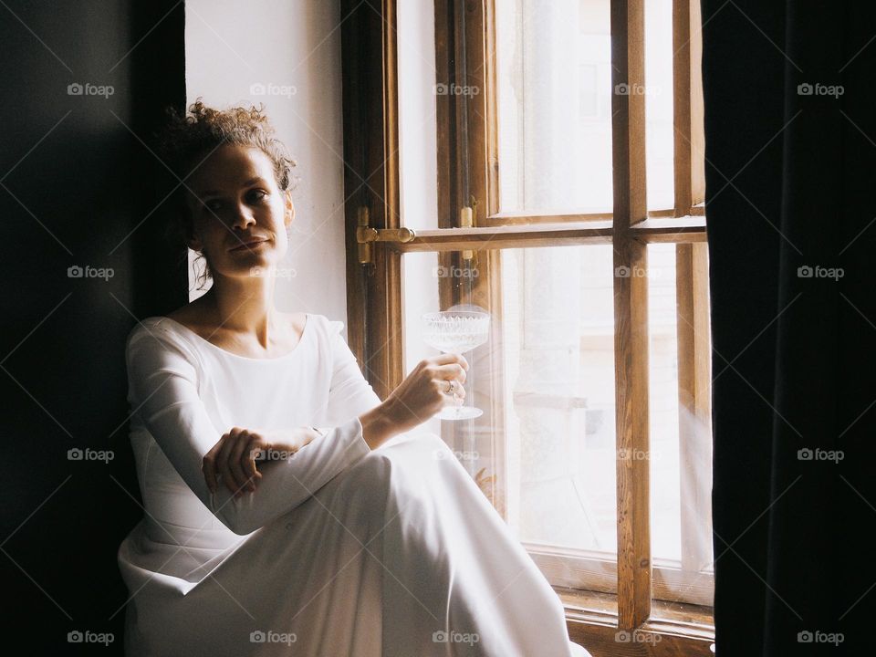 Young beautiful woman with curly hair wearing white dress sits on the window in the bathroom and drinks champagne, portrait of woman, looking away, lifestyle, at home, relax.