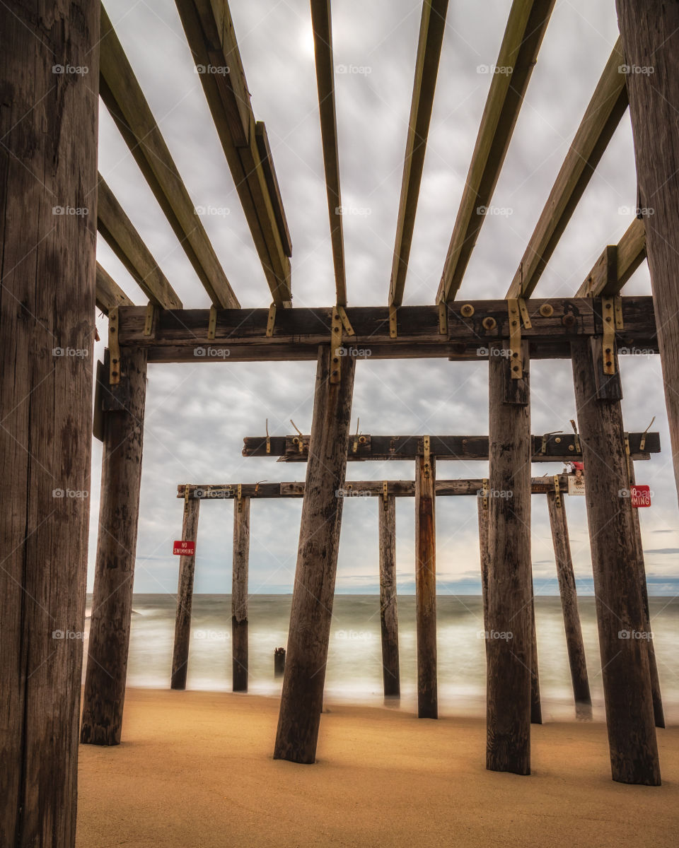 Beams of an old wooden pier leading into the ocean criss crossing to create geometric shapes, framing a moody stormy sky. 