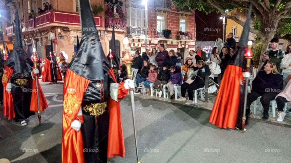 Men wearing Black and Red Gowns, Cloak's and pointed full head Masks, with Staffs in their hands at the Semana Santa Easter Celebrations Night Time Religious Parade through the Main Streets, in the City of Cartagena, Southern Spain. The Crowd Watches