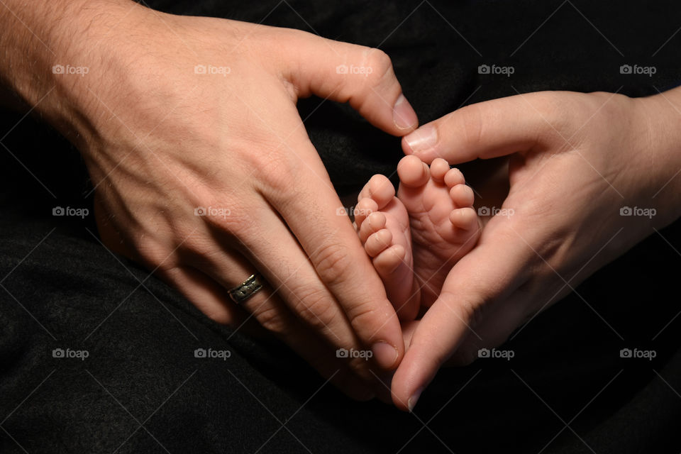Baby’s feet held by mom and dad’s hand shaped like a heart, wedding band visible.