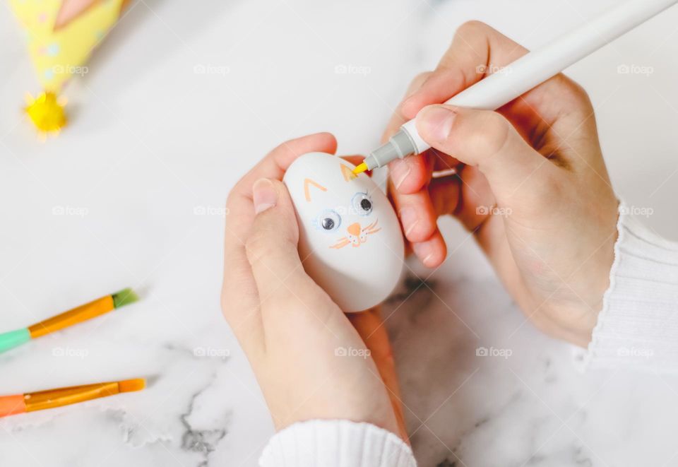 The hands of a caucasian teenager girl draw orange eyebrows with a marker on a white egg with glued eyes and a muzzle of a bunny, preparing a fake for the Easter holiday while sitting at a marble table, close-up side view. Concept diy, crafts, childr