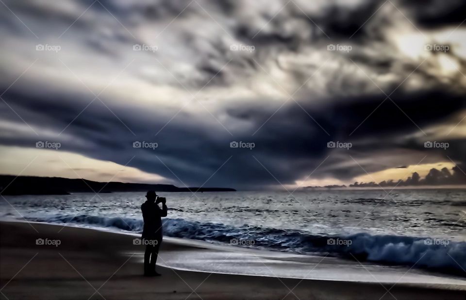 A person takes a photo as dark storm clouds push away the sunset on the beach at Nazaré, Portugal 