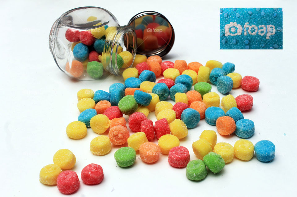Sour chewy gummies flowing out of a small overturned glass jar. The up turned lid reflects the candies. A foap logo & foap name tag made of turquoise & white jelly beans is placed in the top right corner! 😋