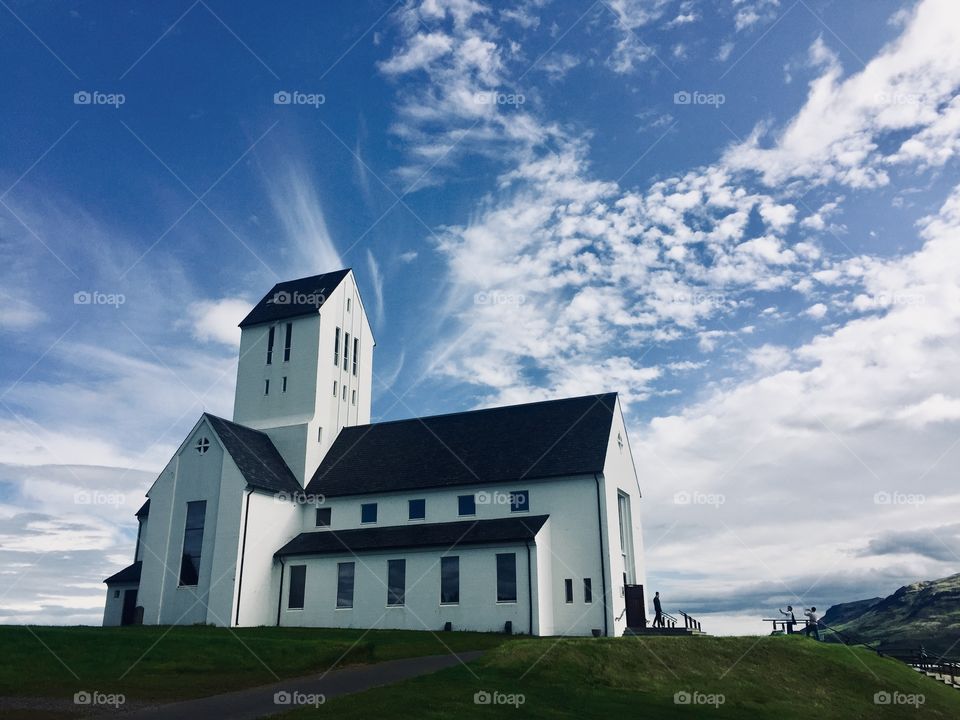 An impressive white church with windows sits on a green hillside in-front of the cloudy, blue sky 