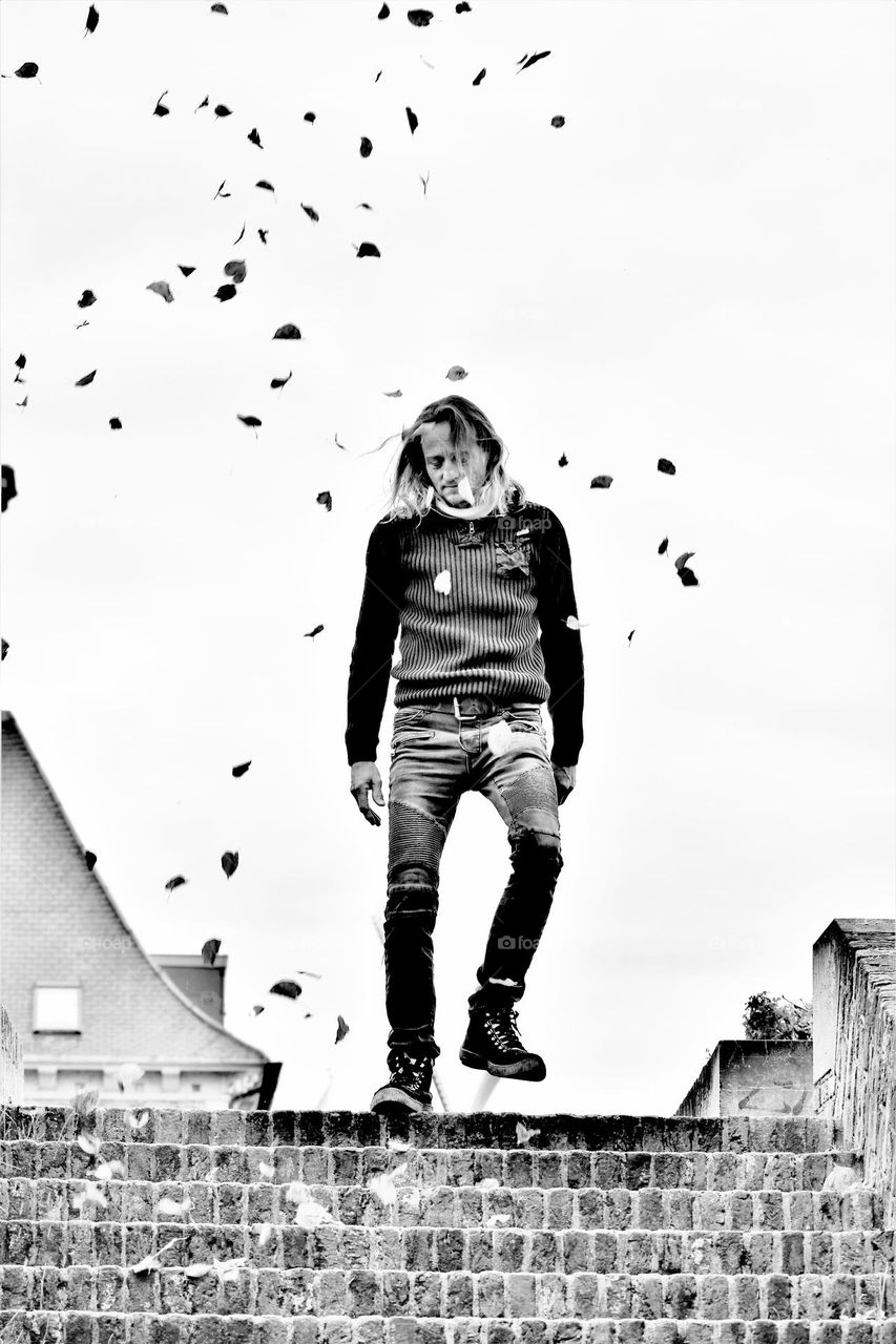 black and white picture from a man with long hair wearing jeans and a sweater in the middle of leafs blowing in the wind